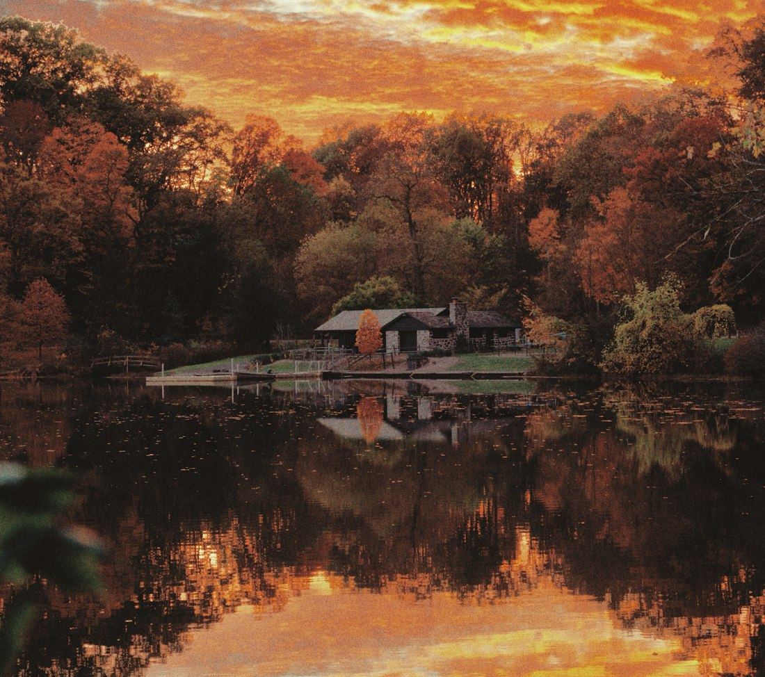 Houten huisje aan een meer bij een vurige zonsondergang, omringd door herfstbos